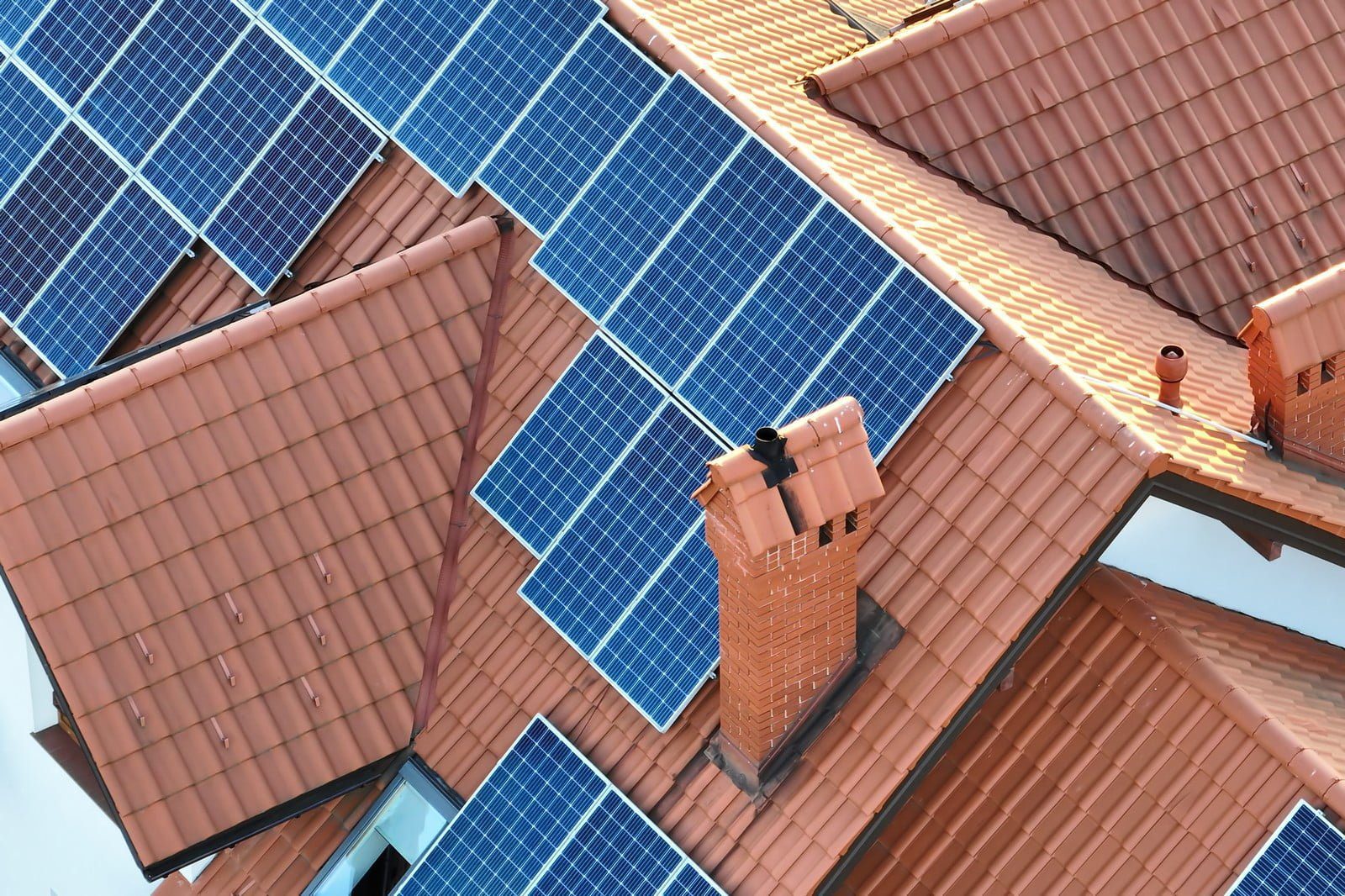The image shows a series of solar panels installed on the roof of a building. The roof has terracotta-coloured tiles, and there are several brick chimneys visible. The solar panels are arranged in multiple arrays, capturing sunlight to convert it into electricity. The installation is typical of residential or small commercial buildings looking to generate renewable energy.