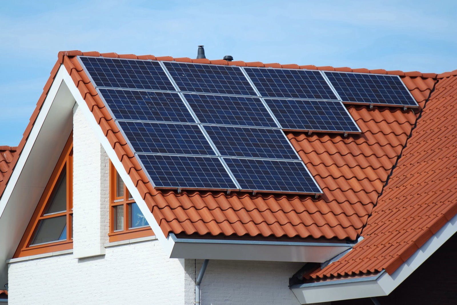 This is an image of a residential house with a number of solar panels installed on its sloped, tiled roof. The solar panels are designed to absorb sunlight and convert it into electrical energy, thereby providing a renewable energy source for the home. The house appears to be two-storied with a white exterior, and it features windows with orange-brown frames that match the roof tile colour. There are no visible people in the image.